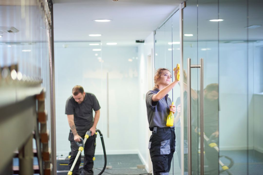 Commercial cleaners wiping glass partitions and vacuuming an office